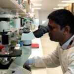 Indra is looking through a microscope. He is wearing a lab coat and gloves.